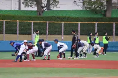 野球場で少年野球の選手たちが、数人のスタッフに見守られながらフィールディング練習をしている様子を捉えた画像です。選手たちはそれぞれ異なるユニフォームを着用し、準備姿勢で身をかがめています。
