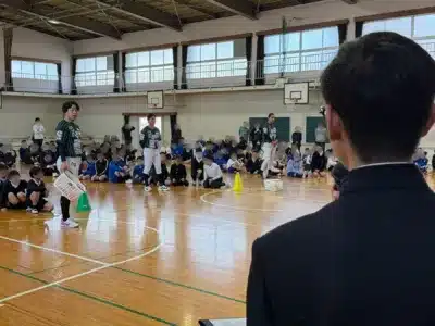 体育館で、中日ドラゴンズのユニフォームを着たプロ野球選手らしき人物が、大勢の小学生や中学生らしき子供たちを前に指導や交流を行っているイベントの様子です。子供たちは床に座って真剣に話を聞いており、スポーツ教室や講演会の一場面と推測されます。