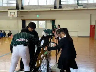 体育館で、ユニフォーム姿と制服姿の男女学生たちが協力してスポーツ用具（バッティングネット）を準備している様子。活気ある学校の活動風景です。