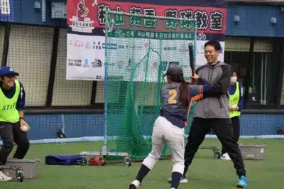 野球教室の様子を捉えた画像です。背番号2の子供がバットを構え、男性コーチが笑顔で指導しています。人工芝のグラウンドで、他のスタッフも見守る中、熱心な練習が行われているようです。背景には野球教室のバナーが見えます。
