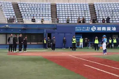 野球場のグラウンドに複数の人々が立っており、奥には空の観客席が広がっています。青い壁には日本語の広告が見え、一部の人は「STAFF」と書かれたベストを着用しています。試合前後の様子やイベントの一場面を捉えた写真です。