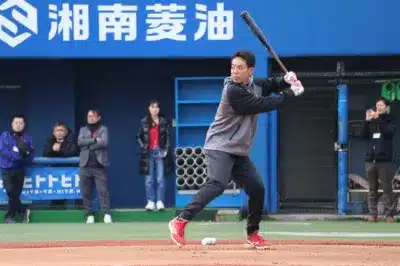 野球場でバッティング練習をする選手の姿を捉えた写真です。青い壁の背景には「湘南菱油」の文字が見え、数名の関係者や観客が練習を見守っています。