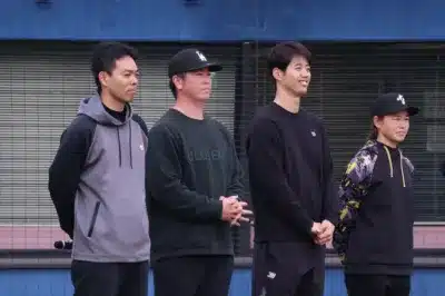 屋外で並んで立つ4人の人物（男性3人、女性1人）が写っています。彼らはカジュアルなスポーツウェアを着用しており、一部の人物はキャップをかぶっています。背景は建物です。