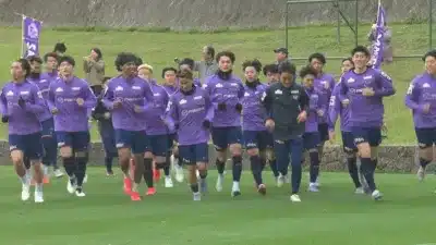 紫色のユニフォームを着たサッカー選手たちが、緑のグラウンドでジョギングをしているトレーニング風景です。背景には数人の観客が見られます。