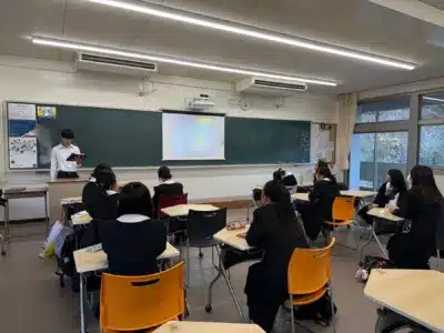 教室で男子生徒がタブレットを手にプレゼンテーションをしており、女子生徒たちが熱心に聞き入っている