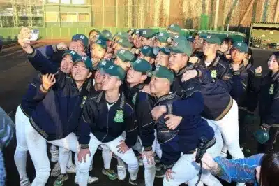 野球部のメンバーらしき大勢の若者たちが、緑のキャップとユニフォームを着用し、屋外で笑顔でセルフィーを撮っている集合写真です。