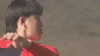 マレットヘアの若い男性が横顔で写っており、赤いスポーツウェアを着て何か活動をしている様子。背景はぼやけている。