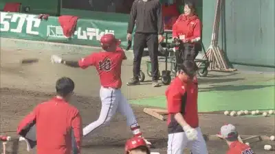 赤いユニフォームを着た野球選手がバットを振り抜き、バットが飛んでいく瞬間を捉えた画像です。グラウンドでは他の選手やスタッフもおり、野球の練習風景が展開されています。背景には「広島」の文字が見えます。
