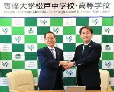 専修大学松戸中学校・高等学校のロゴを背景に、二人の男性が笑顔で握手をしている写真です。学校関係者間の協力や提携を示す公式な場面と推測されます。