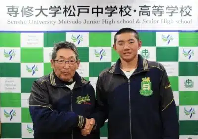 専修大学松戸中学校・高等学校のロゴ入り背景の前で、制服のような上着を着た男性2人が握手を交わしています。年配の男性と若い男性が並んでおり、学校関係のイベントや表彰の場面のようです。