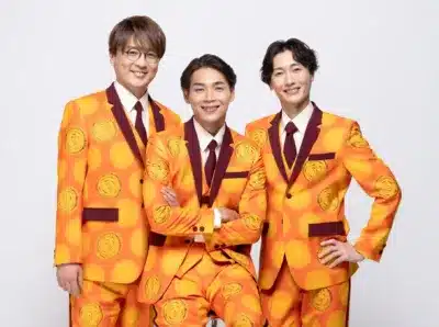 明るいオレンジ色の水玉模様のスーツを着用した3人の男性が、白い背景の前で笑顔でポーズをとっています。彼らは統一感のある衣装で、楽しげな雰囲気を醸し出しています。