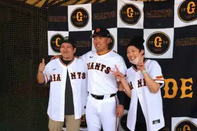 読売ジャイアンツのユニフォームを着たプロ野球選手と2人のファンが、ジャイアンツのロゴが入った背景の前で記念撮影をしています。皆笑顔で、ファンは親指を立てたりピースサインをしたりして楽しんでいる様子です。