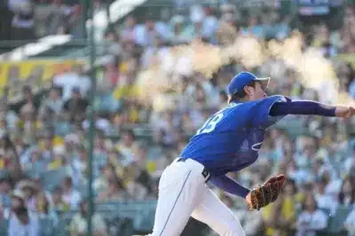 野球の試合で、青いユニフォームを着た投手がボールを投げている瞬間を捉えた写真です。投手の腕からは白い粉が舞い上がり、背景にはぼかされた観客席が広がっています。