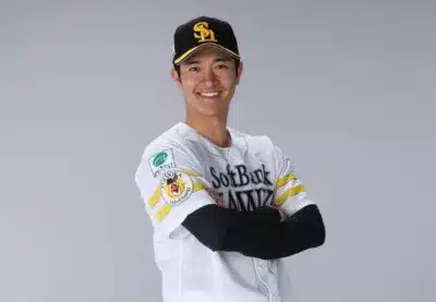 福岡ソフトバンクホークスのユニフォームを着た野球選手が、腕を組み笑顔でポーズをとっています。白いユニフォームにはチームロゴやスポンサー名、福岡の文字が見られます。