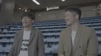 空の観客席を背景に、二人の男性が会話している様子