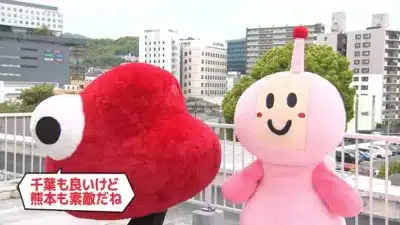屋上に立つ2体のキャラクター（赤とピンク）が都市の風景を背景にしている。赤いキャラクターの吹き出しには「千葉も良いけど熊本も素敵だね」と書かれており、日本の地域に関するメッセージを発信している様子。