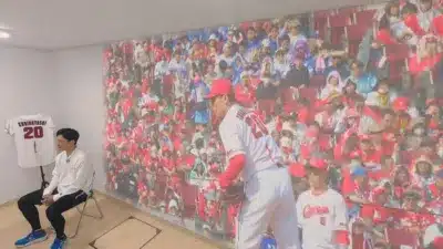 壁面に野球の試合風景が映し出された部屋で、男性が椅子に座り、背番号20のユニフォームが飾られている