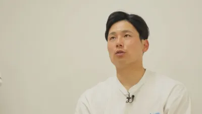 白いTシャツを着た男性が、ラベリアマイクを付けて真剣な表情で話しているか、または聞いている様子