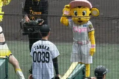 野球場で、阪神タイガースのマスコットがピンクの帽子とユニフォームを着て敬礼しており、背番号「TANAKA 0039」の選手やカメラマンが写っています