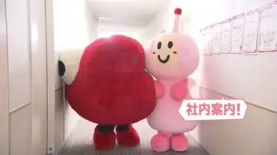 明るい廊下で、赤いキャラクターが壁から顔を出し、ピンクのキャラクターが「社内案内!」と書かれた吹き出しを指している場面です。2体の可愛らしいマスコットが会社を案内しているようです。