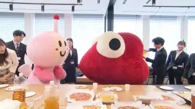 オフィスのような空間で、ピンクと赤の大きなマスコットを中心に、スーツ姿の男女がピザや軽食を囲んで交流している様子。和やかな雰囲気のイベント風景です。