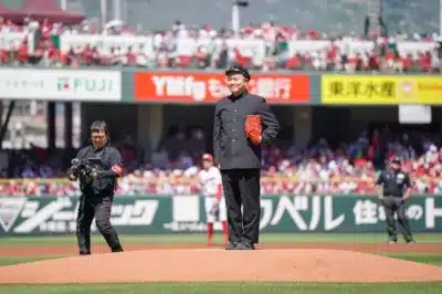 野球場のマウンドで、学生服を着た男性が赤いグローブを持って立っています。始球式のようなイベントが行われているようで、カメラマンや審判もフィールドにいます。観客席には多くの人がおり、天気の良い日中の様子です。