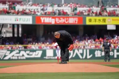 野球場のマウンド上で、学生服のような黒い衣装と赤いグローブを身につけた男性が深くお辞儀をしている。背景には多くの観客と「もみじ銀行」「東洋水産」などの看板が見え、始球式の様子を捉えたものと推測される。