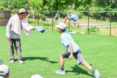 人工芝のグラウンドで、大人の男性と少年がキャッチボールをしている様子です。男性は指導しているように見え、少年はボールを投げようとしています。