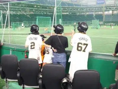 野球場で練習を観戦する人々が写っています。吉川2番、山瀬67番、大勢15番のユニフォームを着たファンがヘルメットを着用し、バックネット越しにグラウンドを見つめています。親子連れで野球を楽しんでいる様子が伝わります。