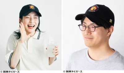 阪神タイガースのロゴ入りキャップを被った男女が写っています。女性は笑顔でコップを持ち、男性は眼鏡をかけ微笑んでおり、タイガースのグッズをアピールしているようです。