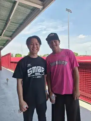 屋外のスポーツ施設らしき場所で、2人の男性が笑顔で並んで立っている写真