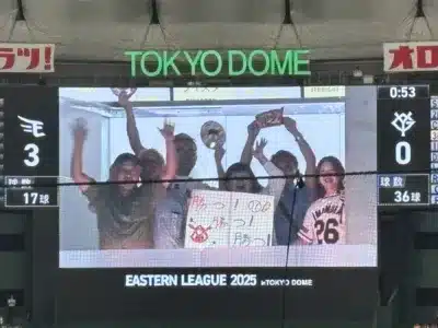 東京ドームの大型ビジョンに映し出された野球の試合風景です。観客が「勝つ!」と書かれた応援ボードを持ち、熱心に応援している様子が映し出されています。スコアボードには「EASTERN LEAGUE 2025」と表示され、試合情報も見えます。