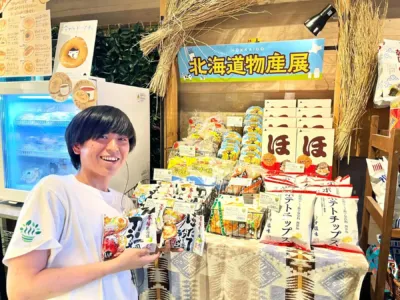 北海道物産展のブースで、笑顔の若い男性が北海道の特産品を手にしている。