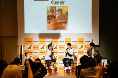 タイガーエア台湾のイベントで、ステージ上では複数の男性がマイクを持って話しており、観客がそれを見守っています。背景のスクリーンには「tigerair」のロゴと、巨大なフルーツデザートを囲む2人の男性の写真が映し出されています。
