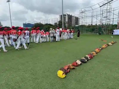 人工芝の野球場で、赤と白のユニフォームを着た少年野球の選手たちが集まり、練習またはアカデミーのイベントに参加している