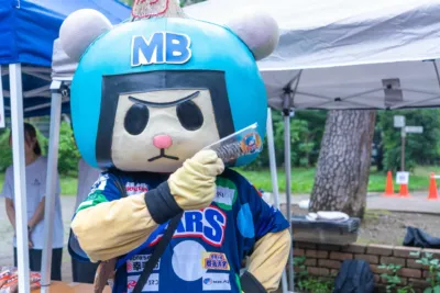 青いヘルメットをかぶったマスコットキャラクターが、屋外イベント会場で何かを手に持っています。