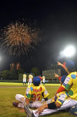 夜の野球場で、選手たちがグラウンドに座ったり立ったりして夜空に打ち上がる壮大な花火を見上げている。