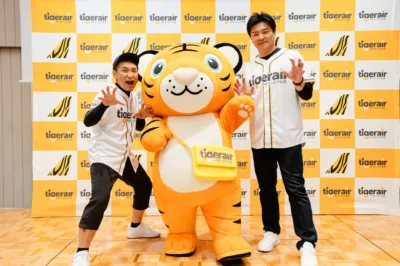 タイガーエア台湾のロゴ入り背景の前で、野球ユニフォームを着た男性2名と可愛らしいトラのキャラクターが笑顔でポーズをとっています。