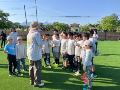 青空の下、緑のグラウンドでコーチが子供たちに何かを説明しているスポーツ指導の場面です。多くの子供たちが集まり、熱心に耳を傾けています。
