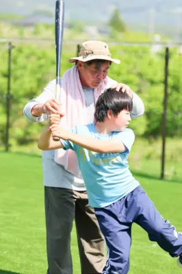 晴れた屋外のグラウンドで、大人の男性が少年に対し野球のバッティングを指導している様子。少年は真剣な表情でバットを構えている。