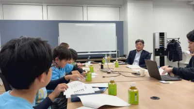 会議室で子供たちと大人が集まり、学習やディスカッションを行っている様子。机の上には資料やノートパソコン、伊右衛門のペットボトルが並び、大人も子供も熱心に取り組んでいるワークショップのような光景です。