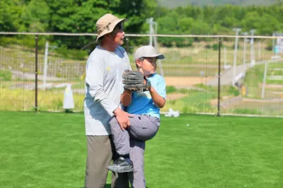 晴れた屋外の人工芝のグラウンドで、大人の男性が少年に野球のピッチングフォームを指導している様子。少年はグローブをはめ、男性のサポートを受けながら投球動作を確認している。