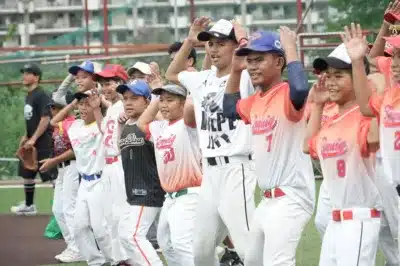 野球のユニフォームを着た少年たちが屋外のグラウンドに一列に並び、笑顔で手を上げている様子