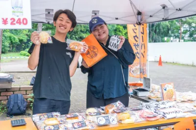 屋外の販売ブースで、笑顔の男女が「小麦の奴隷」のパンを販売している。
