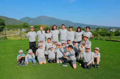 明るい日差しの下、大人と子供たちが揃いのグレーのユニフォームと帽子を身につけ、人工芝の広場で集合写真を撮っています。背景には山々が広がり、参加者たちは楽しそうにポーズを取っています。犬も一緒に写っており、一体感が感じられる屋外イベントの様子です。