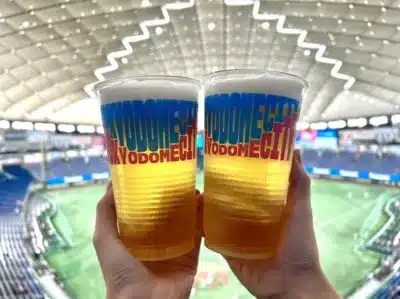 東京ドームの野球場で、2つのビールカップを掲げている様子。カップには「TOKYODOME CITY」のロゴがあり、野球観戦中の楽しい雰囲気が伝わってきます。