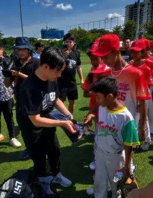 屋外のスポーツフィールドで、大人が野球ユニフォーム姿の子供たちに何かを手渡している