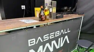 「BASEBALL MANIA」のロゴが描かれたカウンターに、ウイスキーや焼酎などの酒瓶、テレビ、スピーカーが並べられています