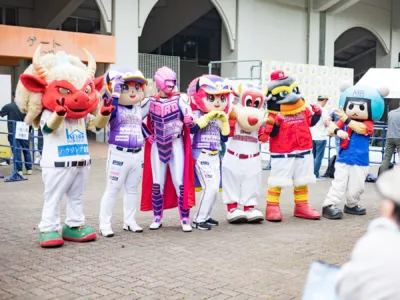 野球場のゲート前で、様々なプロ野球チームやイベントのマスコットキャラクターたちが集合し、ポーズをとっている