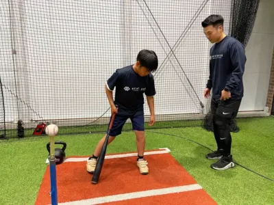 室内練習場でティーバッティングをする子供とコーチ